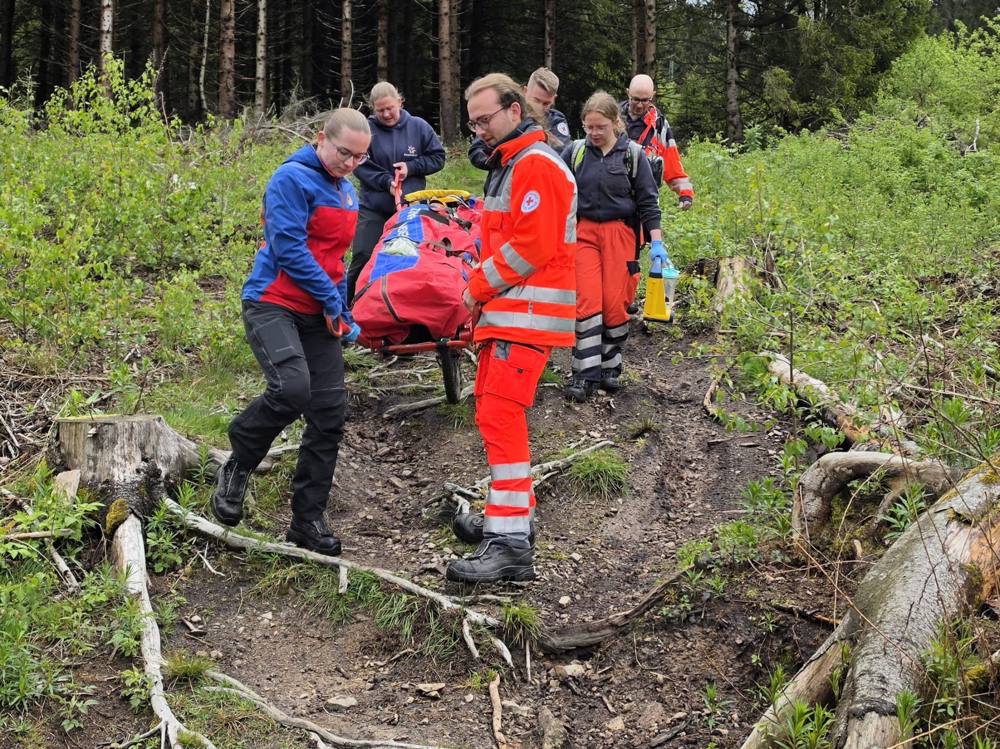 Einsatzkräfte der Bergwacht und des Rettungsdienstes transportieren eine Gebirgstrage über einen matschigen Waldweg